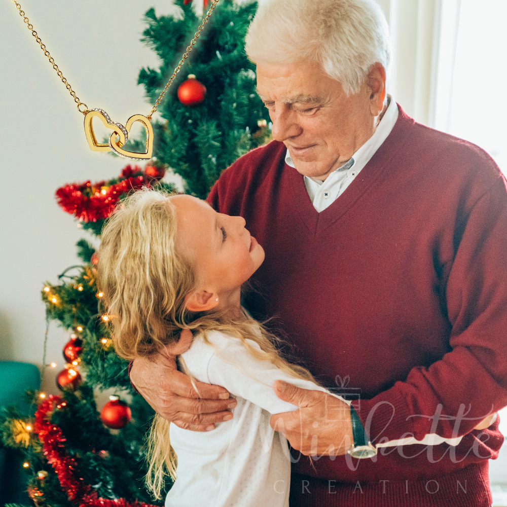 Grandpa and granddaughter embracing in front of a decorated Christmas tree, with a necklace featuring a heart-shaped pendant.