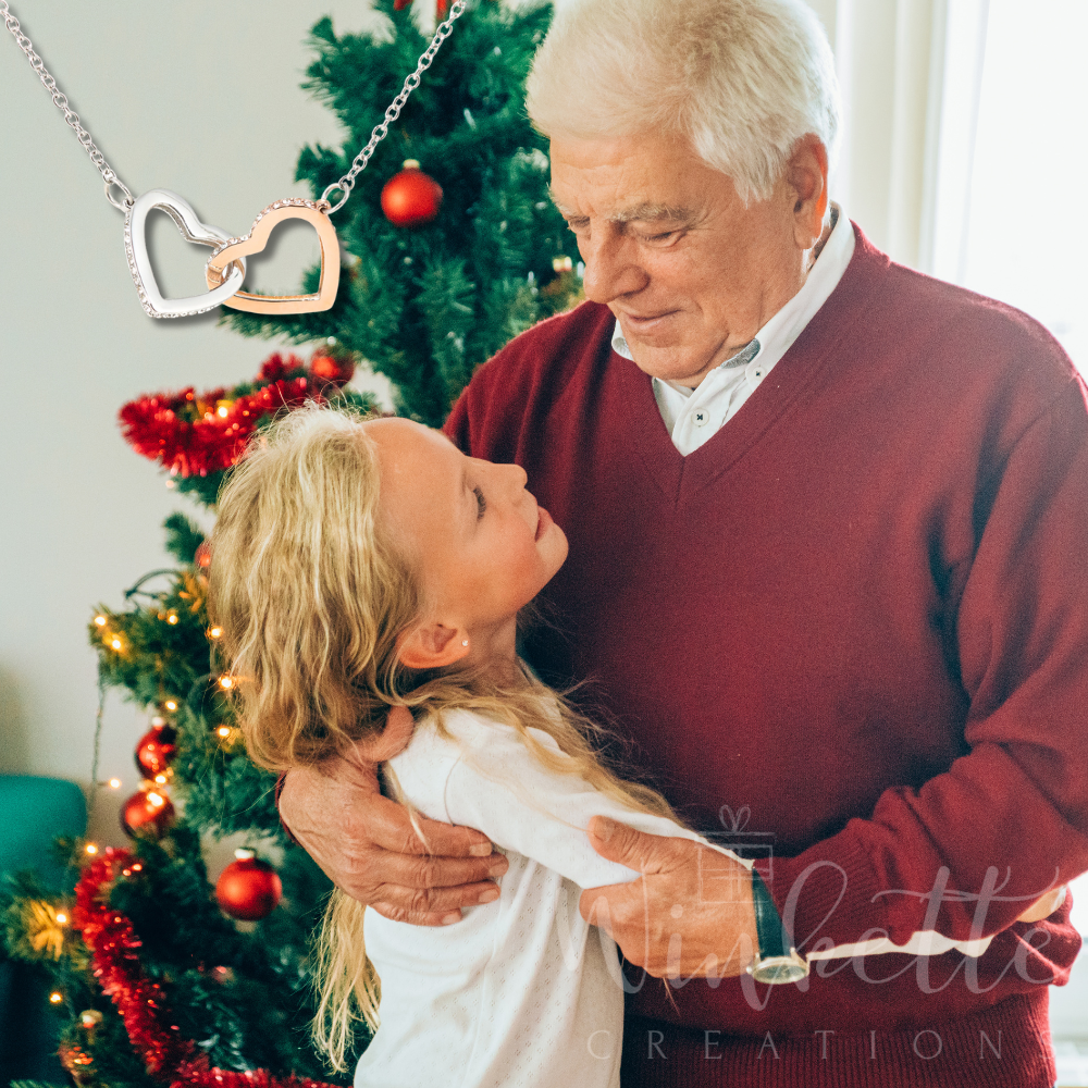 grandpa and granddaughter embracing in front of a decorated Christmas tree, with a necklace featuring a heart-shaped pendant.