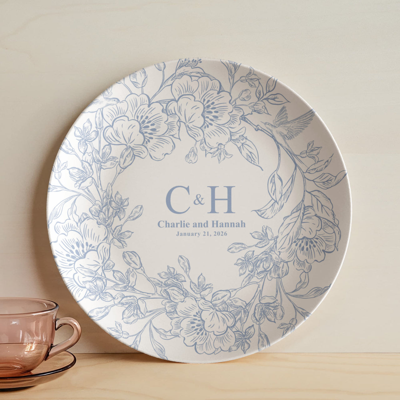 A round white polymer plate with a delicate blue floral border and personalized monogram text reading 'C&H Charlie and Hannah January 21, 2026', styled on a light wood surface next to a translucent pink glass teacup.