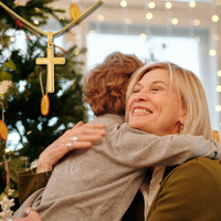 Thumbnail for grandson and grandma hugging in front of a decorated Christmas tree with lights in the background.
