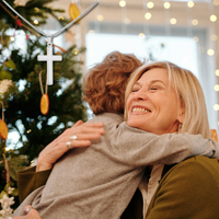 Thumbnail for grandson and grandma hugging in front of a decorated Christmas tree with lights in the background.