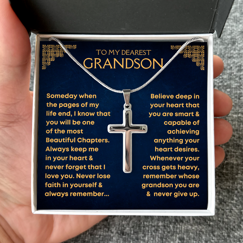 A silver cross pendant necklace displayed inside a gift box with a navy blue message card addressed 'TO MY DEAREST GRANDSON,' held in a hand.