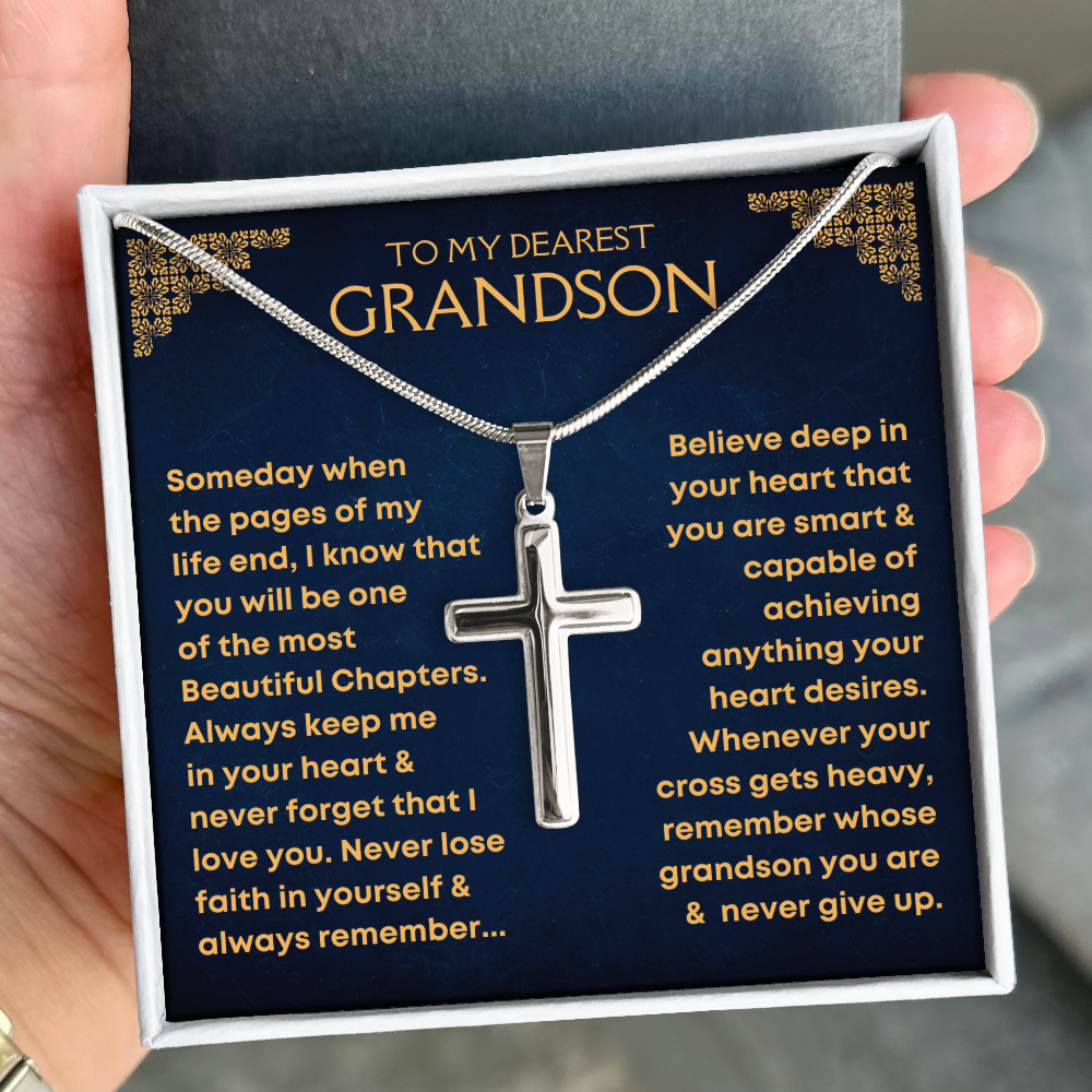 A silver cross pendant necklace presented in a gift box with a dark blue message card addressed to a grandson, held in a hand.