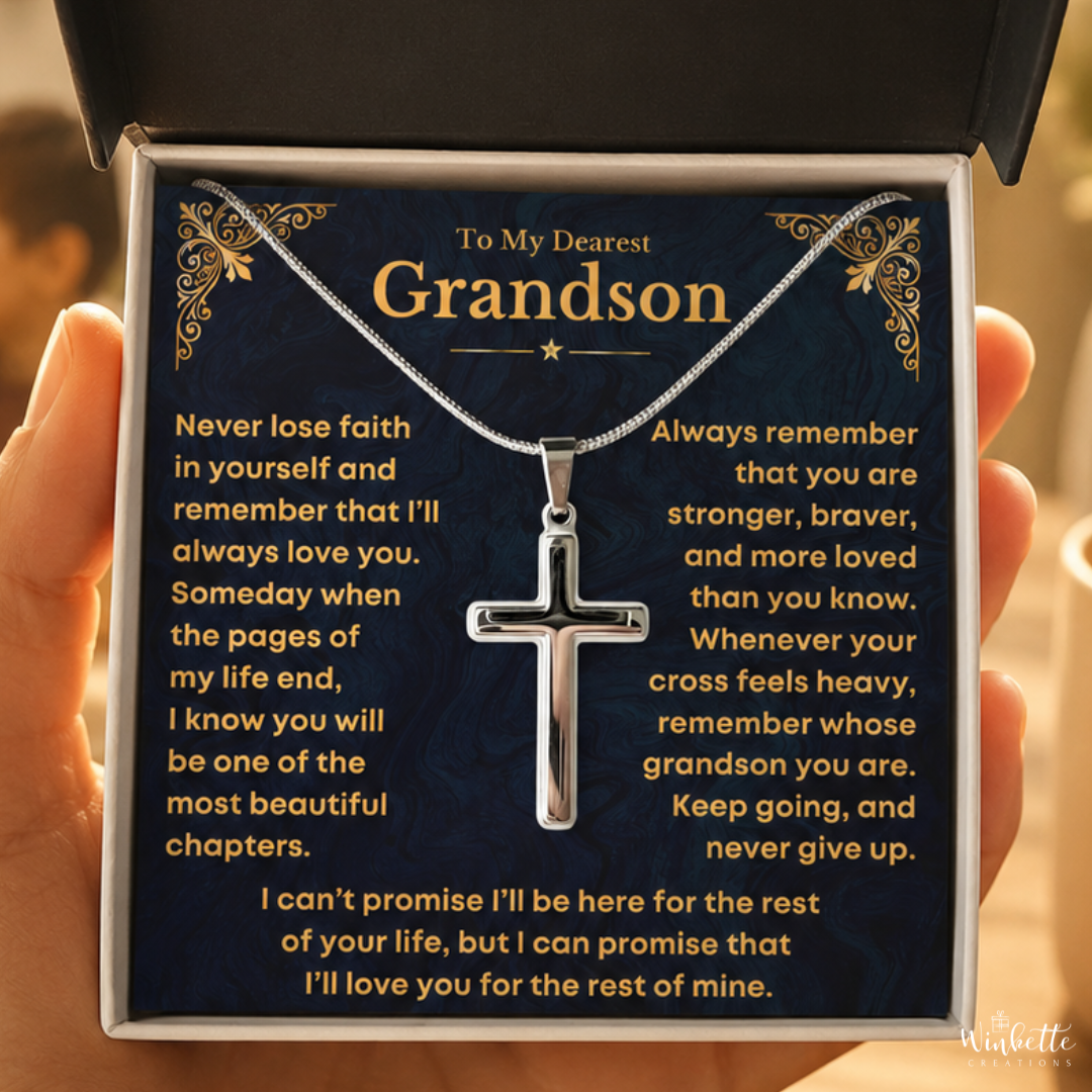A silver cross pendant necklace displayed on a dark blue message card inside a gift box, held in a hand. The card is addressed 'To My Dearest Grandson' and features gold decorative corner details and yellow text with a sentimental message from grandma grandpa