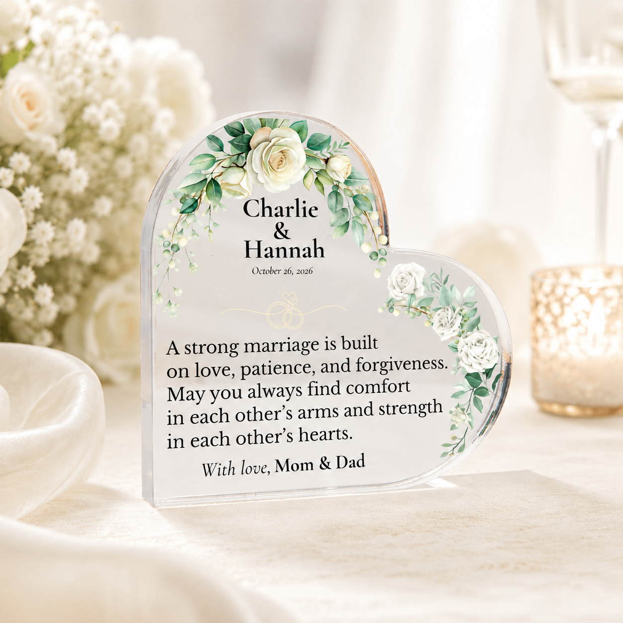 A heart-shaped clear acrylic keepsake plaque with floral green and white illustrations, personalized with the names 'Charlie & Hannah' and a memorial message, displayed on a table with soft-focus wedding decor in the background.