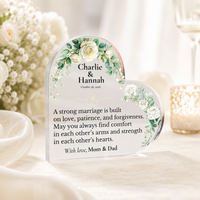 Thumbnail for A heart-shaped clear acrylic keepsake plaque with floral green and white illustrations, personalized with the names 'Charlie & Hannah' and a memorial message, displayed on a table with soft-focus wedding decor in the background.