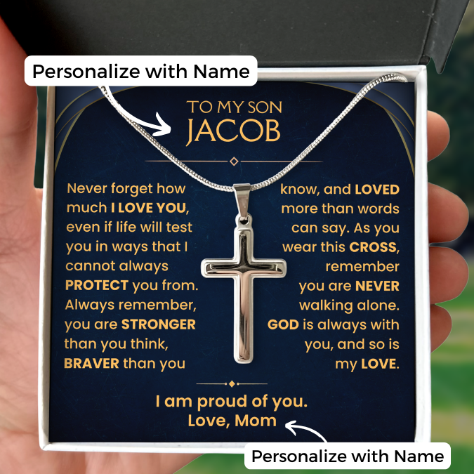 A silver cross pendant necklace displayed on a personalized dark blue gift card inside a white box, held by a hand. The card features gold text with a message for 'my son Jacob' and white 'Personalize with Name' labels.
