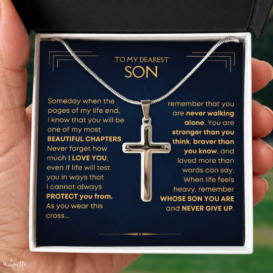 A silver cross pendant necklace displayed inside an open gift box with a dark navy message card addressed to a son, held in a person's hand.