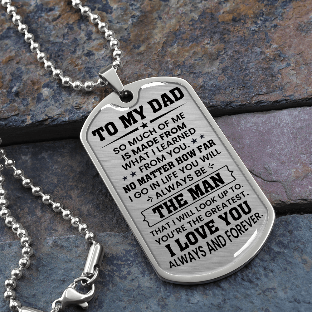 A silver dog tag necklace with the engraving 'TO MY DAD' and a message in bold letters, attached to a military-style chain placed on a textured background.