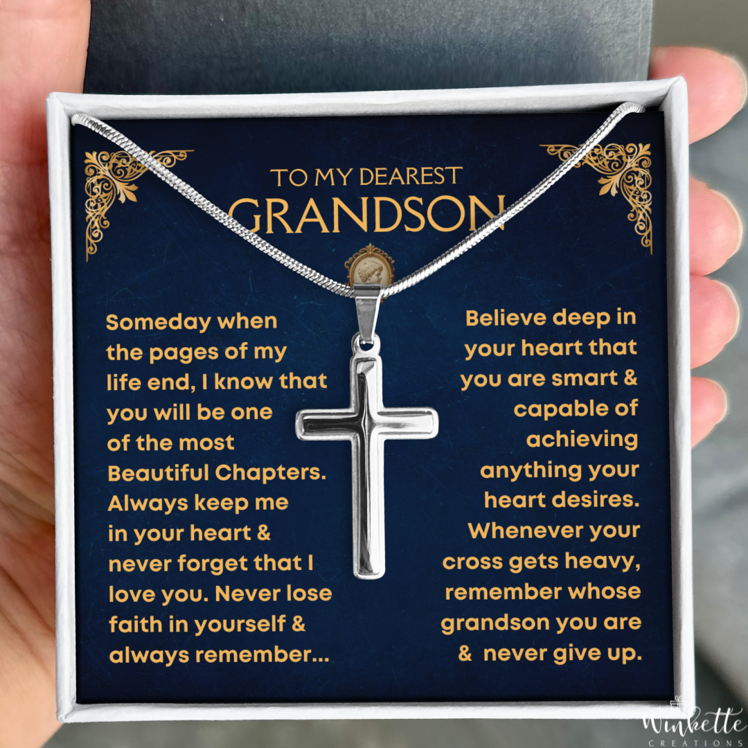 A silver cross pendant necklace presented in a gift box with a dark blue message card addressed to a grandson. A hand is holding the box open, and the necklace is displayed against the card.