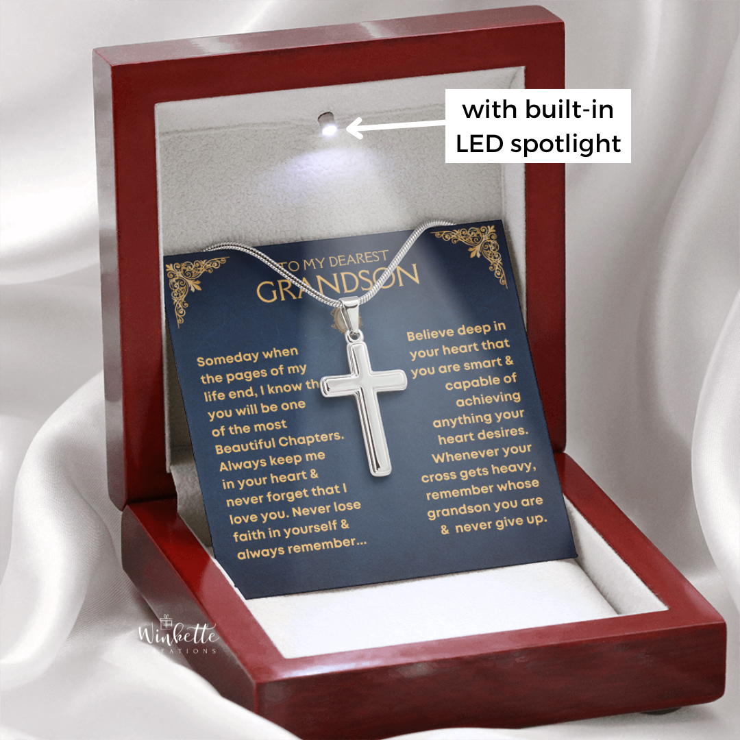 A silver cross necklace presented inside a wooden keepsake box with a built-in LED spotlight. The silver cross is displayed over a message card for a 'dearest grandson.